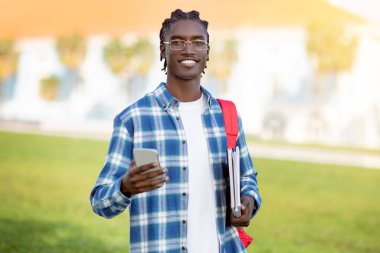 Ekose gömlekli ve gözlüklü siyahi öğrenci cep telefonunu açık havada kameraya gülümserken aynı zamanda üniversitede çağdaş gençlere yönelik teknoloji ve eğitimin karışımını gösteriyor.
