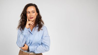 Kıvırcık saçlı, mavi tişörtlü ve bej pantolonlu, özgüvenli beyaz bir kadın, düşünceli, çenesi dinlenmiş, profesyonellik ve duruş dolu, panorama