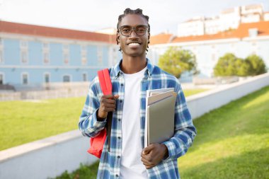 Kampüs binasına karşı poz verirken çalışma kitabı, bilgisayar ve sırt çantası tutan neşeli siyahi öğrenci, kameraya gülümsüyor ve yurtdışında eğitime hazır. Öğrencilik yaşam tarzı
