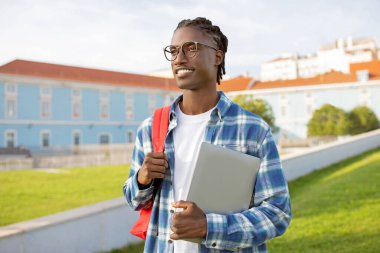 Mesafe öğrenme. Hevesli Afrikalı Amerikalı Öğrenci, sırt çantasıyla kampüs parkındaki üniversite binasında dizüstü bilgisayarı tutuyor ve gülümseyerek uzaklara bakıyor. E-Öğrenme Teknikleri
