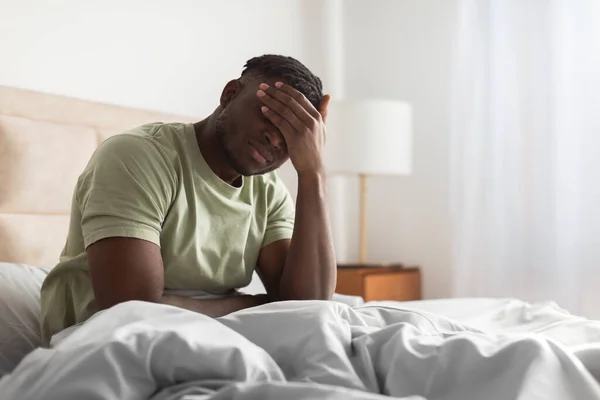Sleepy tired black man in bed covering face with hand, struggling from insomnia and morning headaches, sitting at home bedroom interior. Free space for text. Male healthcare concept
