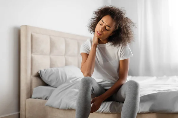 Tired african american millennial woman touching aching neck, suffering from pain after uncomfortable sleep, sitting on bed and massaging neck in modern bedroom at home. Osteochondrosis