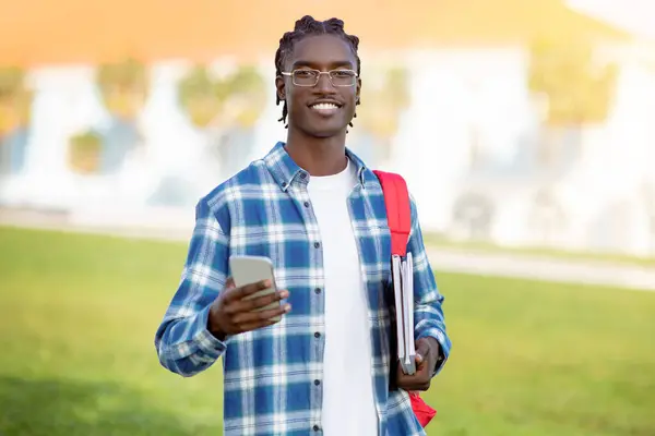 Black student guy in plaid shirt and eyeglasses uses cellphone standing outdoors, smiling to camera while showcasing the blend of technology and education for contemporary youth in university