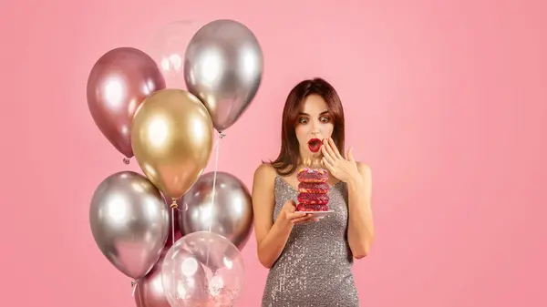 Astonished young caucasian woman in a sparkling dress looking at a stack of colorful donuts with candles, surrounded by helium balloons, celebrating a fun and festive occasion