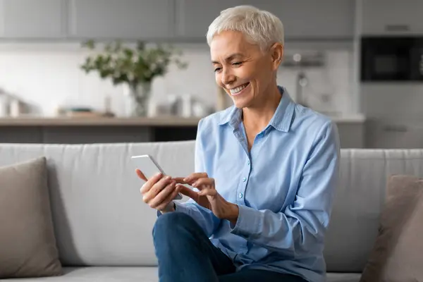 Mobile Technology. Happy Mature Woman Using Smartphone Gadget And Applications, Reading Online News, Texting And Communicating On Couch, Sitting In Cozy Living Room At Home Interior
