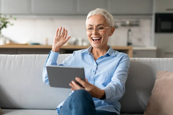Virtual Communication. Happy Mature Woman In Eyeglasses Makes Video Call Using Digital Tablet Computer, Gesturing Hello Greeting To Webcam, Communicating Online Sitting On Couch At Home