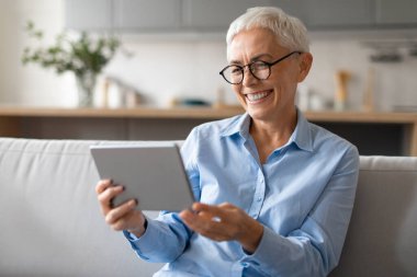 Yaşlı bayan dijital tablet kullanıyor. İnternette kitap okuyor. Evdeki koltukta oturuyor, gözlük takıyor. Mutlu Kadın Çevrimiçi Eğleniyor Taşınabilir Bilgisayar ile, Film İzliyor ve İnternet Sörfü Yapıyor