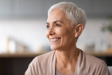 Yaşlanmaz güzellik. Kısa saçlı, şık gri bir kadının portresi gülümseyen, iç mekâna bakan, günlük tarz bej giysiler giyen. Mutlu, zarif, yaşlı bir kadının vesikalığı.