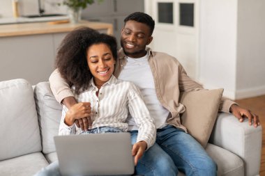 Mutlu Afro-Amerikan çift dizüstü bilgisayar kullanarak video veya film seyrediyor, web sitelerine bakıyor ve bilgisayar başında eğleniyor, oturma odasındaki kanepede dinleniyor. İnsanlar ve internet teknolojileri