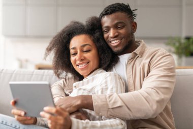 Gülümseyen siyah bir çift tablet tablet üzerinde sosyal medyada geziniyor, evlerindeki koltukta rahatça oturuyor ve kucaklaşıyorlar, karı koca interneti keşfederken arkadan sarılıyorlar.