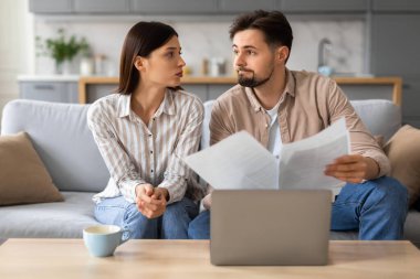 Endişeli genç çift evrakları ciddi ifadelerle inceliyor, masadaki, evdeki dizüstü bilgisayarlar, finansal planlamayı öneriyor.