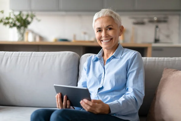 Mature Woman With Stylish Haircut Posing With Digital Tablet Computer, Sitting On Couch And Smiling To Camera While Relaxing With Gadget, Browsing Internet At Home Interior, Empty Space