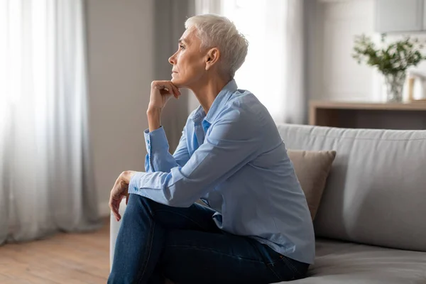 Depression, Mental Health Issue. Depressed senior woman touching chin sitting on couch at home, looking aside with unhappy thoughtful expression, having problem in retirement life