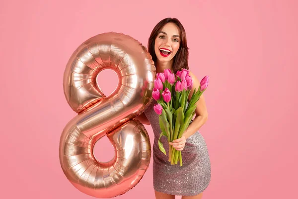 Radiant glad millennial caucasian woman in a sparkling dress joyfully holds a bouquet of pink tulips and a number 8 balloon, celebrating a special occasion on a pink backdrop