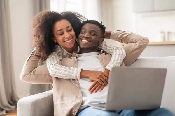 Black Family Couple Hugging While Watching Movie On Laptop Online, Enjoying Weekend Together At Home, Sitting On Sofa In Cozy Modern Living Room. Happy Relationships, Leisure Concept