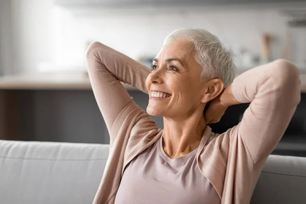 Relaxed senior lady enjoying quiet pause comfortably leaning back on her couch, resting holding hands behind head, embodying relaxation and weekend comfort in modern living room interior