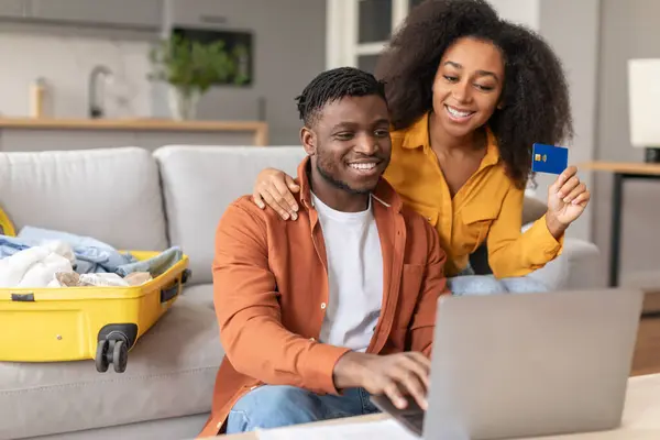 Happy black family couple using laptop computer and credit card buying travel tickets, making payment transaction while booking hotel sitting in living room interior with unpacked suitcase