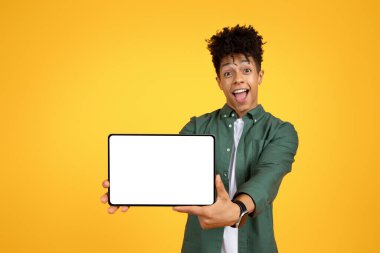 Heyecanlı, şık, yakışıklı, Afro-Amerikalı genç adam beyaz ekranlı dijital tablet gösteriyor ve güzel mobil uygulama, online uygulama, sarı stüdyo arka planı, kopyalama alanı öneriyor.