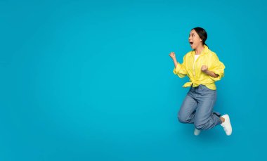 Parlak sarı bluz ve kot pantolon giymiş, havada zıplayan mutlu Çinli genç bir kadın turkuaz mavi bir arka plana karşı neşe ve zafer ifade ediyor.