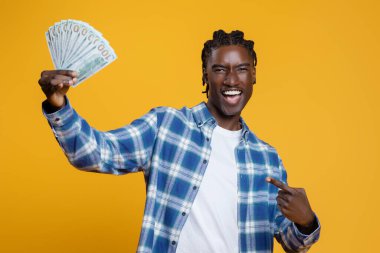 Elinde dolar nakit para tutan ve işaret eden mutlu siyah adam, para banknotlarını gösteren neşeli bir adam, sarı stüdyonun arka planında duruyor, finansal başarıyı ve kârı kutluyor.