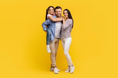 Genç kızlarıyla birlikte sarı arka planda poz veren, üç kişilik beyaz bir aileyi seven ve kameraya gülümseyen mutlu ve genç ebeveynler, fotokopi uzayı ile uzun bir çekim yapıyorlar.