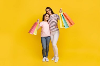 Aile alışverişi. Mutlu genç anne ve genç kız bir sürü parlak alışveriş çantasıyla poz veriyor, neşeli anne ve tatlı kız çocuğu sarılıp, sarı arka planda duruyor, boşluğu kopyalıyor