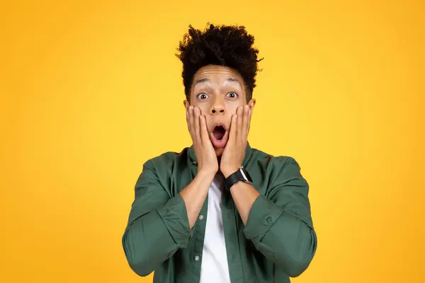 Amazing offer, deal, shocking news. Surprised young african american guy with stylish hair looking at camera, touching face and grimacing, isolated on yellow studio background