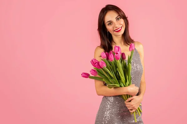 Charming young caucasian woman with a beaming smile, clutching a vibrant bouquet of pink tulips, dressed in a sparkling sequined dress against a soft pink backdrop, studio