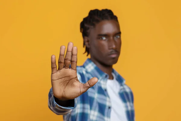 Young black man gesturing STOP with open palm on yellow studio background. Displeased African American guy saying NO, showing rejection gesture with hand, expressing his negative attitude
