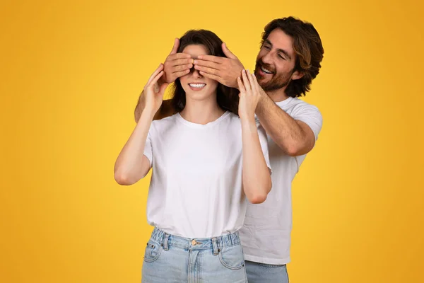 Smiling european millennial man surprises a woman by covering her eyes from behind with his hands, both dressed in white t-shirts, creating a playful moment on a yellow background