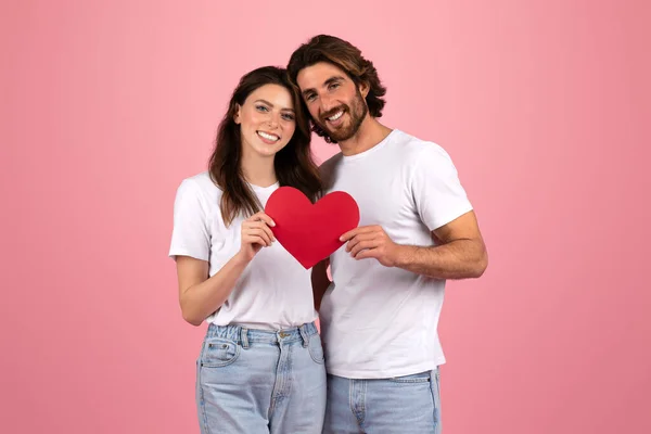 Happy european couple in white t-shirts and jeans holding a red paper heart together, looking at each other with affection, symbolizing love and togetherness on a pink background