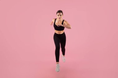 Kameraya doğru koşan kararlı bayan atlet, yüksek enerjili antrenman yapan genç kadın, yumuşak pembe stüdyo arka planına karşı antrenman, fotokopi alanı ile tam boy çekim.