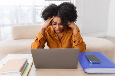 Mutsuz, genç, kıvırcık, Afro-Amerikan kadın öğrenci müdürü oturma odasındaki bilgisayarda stres yaşıyor. İş çalışması ev ofisindeki sorunlar, son teslim tarihi, hata, aşırı çalışma