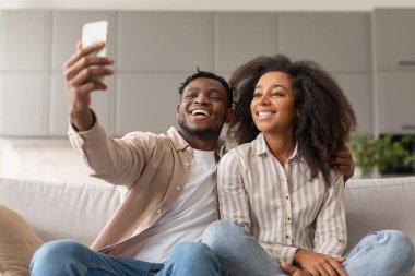 Genç mutlu siyahi eşler cep telefonuyla arayan videoları seven erkek, karısının birlikte poz verdiği, akıllı telefonla eğlenen ve iletişim kuran, evdeki kanepede oturan karısını kucaklıyor.