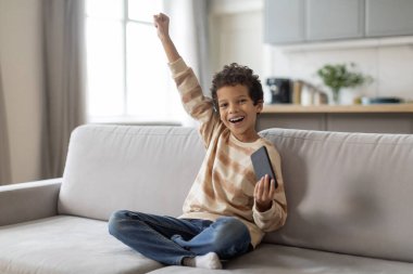 Küçük siyah çocuk evde akıllı telefonuyla oyunlar oynuyor ve başarıyı kutluyor, neşeli çocuk yumruğunu kaldırıyor ve evet diyor, oturma odasında otururken online kazanmanın tadını çıkarıyor.