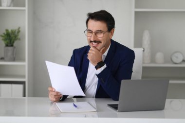 Masada dizüstü bilgisayarlı, düşünceli, modern ofis içi sözleşmeleri okuyan, takım elbiseli, mutlu beyaz iş adamı. Evrak işleri, iş anlaşmaları ve başlangıç için fikir oluşturma