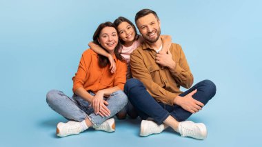 Mutlu Aile. Mavi Stüdyo 'nun arka planında oturan karısını ve kızını kucaklayan neşeli beyaz adam. Ebeveynlerin ve kameraya gülümseyen kızın uzun pozu. Birlikte poz veriyorlar. Panorama