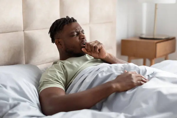Sick Black Young Man Coughing In Fist Suffering Pneumonia Symptom Lying In Bed At Home Bedroom. Ill African American Guy Having Inflamed Lungs Symptoms During Influenza Disease