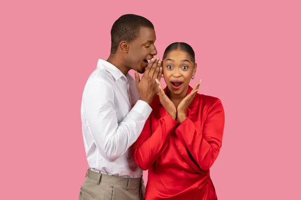 Black man in white shirt whispering into womans ear, her expression of delighted shock, hands on face, against playful pink background