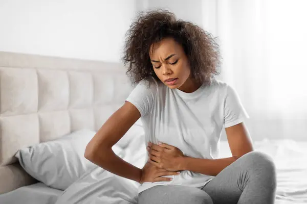 Unhappy black young lady clutches her abdomen sitting on bed, suffering from discomfort having menstrual cramps or digestive issues. Stomachache and female health problems