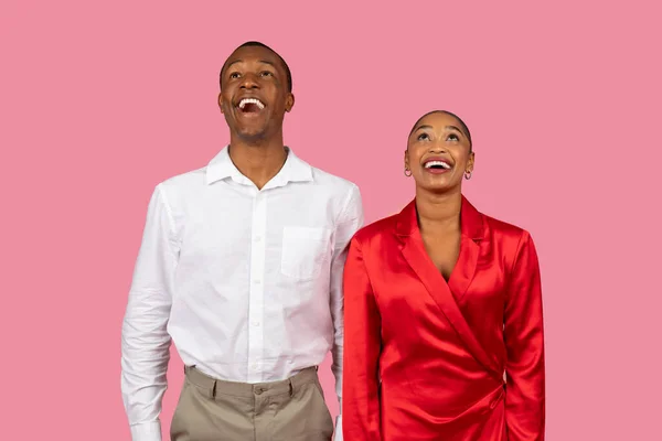 Exuberant surprised black man and woman in stylish white shirt and red dress looking upwards at free space with joy on playful pink background