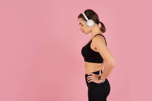 Side profile of determined young female athlete wearing wireless headphones, millennial woman in sportswear listening to music or coaching during fitness routine, standing on pink studio background