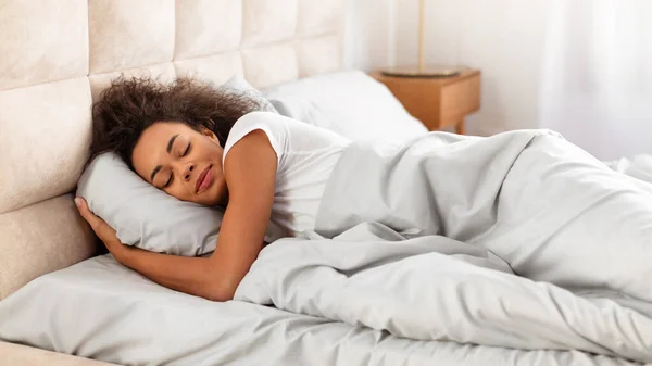 Sleeping African American Young Lady Lying Resting Head On Pillow In Modern Bedroom Interior, Enjoying Recreation Routine, Napping Covered With Blanket In Bed Indoors. Panorama