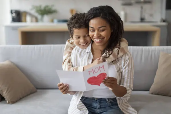 Smiling black mother reading handmade heart card from her cheerful son, loving male kid greeting mom with birthday or mothers day, creating heartwarming moment at home, copy space