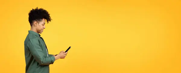 Stylish handsome young black guy wearing green shirt using cell phone, isolated on yellow studio background, panorama with blank copy space for mobile app advertisement