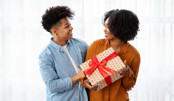 A joyful african american young couple shares a special moment, exchanging a beautifully wrapped gift with a red ribbon, their faces lit up with smiles in a light, airy room