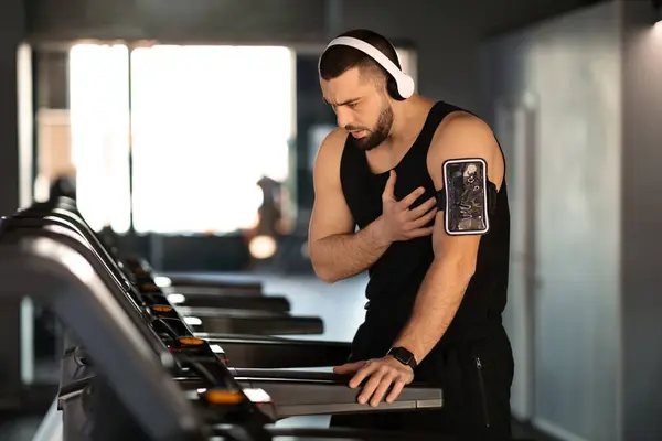 Distressed man experiencing sudden heartache while working out on treadmill, young male athlete showing signs of discomfort and pain during training in gym, touching chest area, copy space
