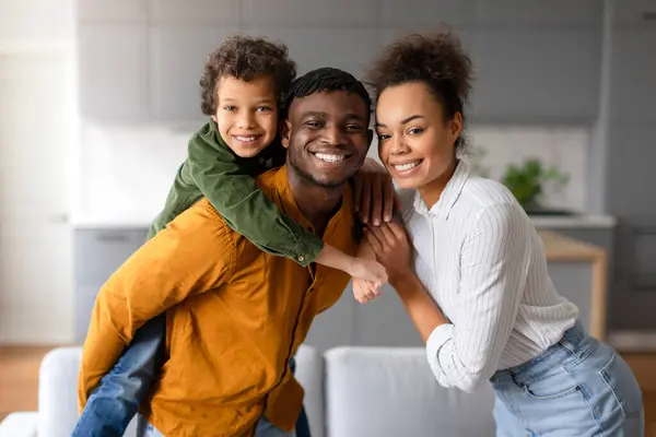 若い男の子が父親の背中にピギーバックに乗って楽しむ陽気な黒人の家族の瞬間は 母親が愛情を持って彼らをサポートし 家庭内でポーズ — ストック写真