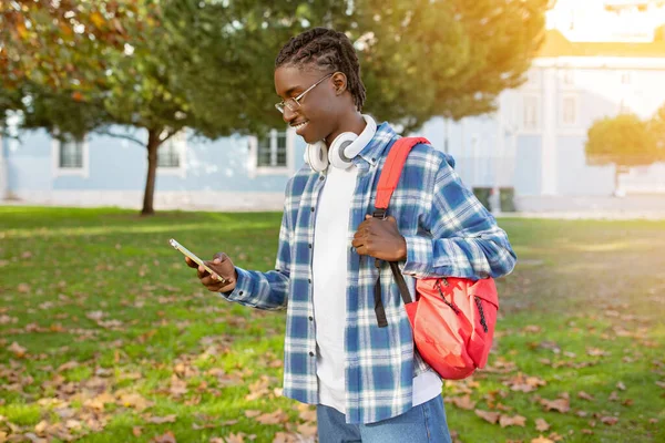 Smiling black student guy in glasses browsing on his phone with headphones ready for music, enjoys sunny day outdoors walking with backpack at university campus park