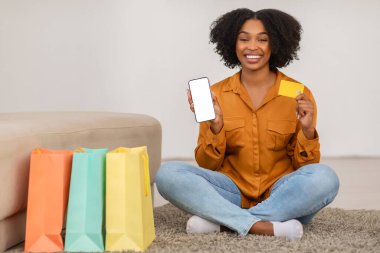 Bin yıllık Afro-Amerikan kıvırcık kadın çantalarla yerde oturuyor, oturma odasının içinde boş ekranlı kredi kartı ve telefon gösteriyor. Çevrimiçi alışveriş tavsiyesi, evde satış